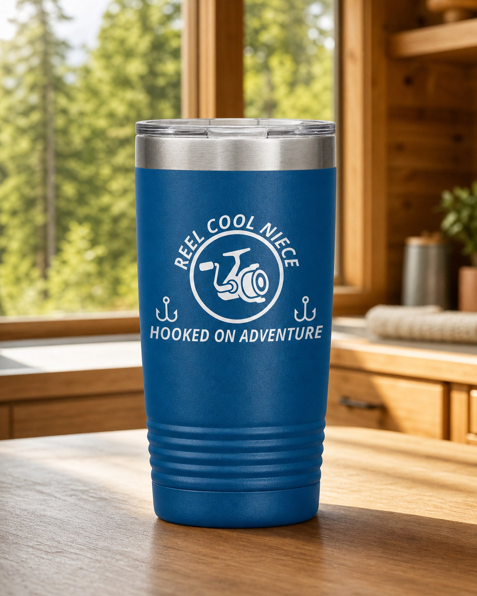 Blue 20-ounce REEL COOL NIECE engraved tumbler on a bright wood cabin counter beside a window overlooking green forest.