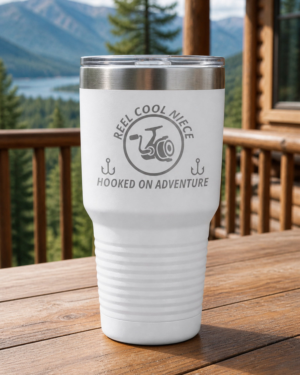 White 30-ounce REEL COOL NIECE engraved tumbler on a sturdy cabin porch table overlooking forested mountains.