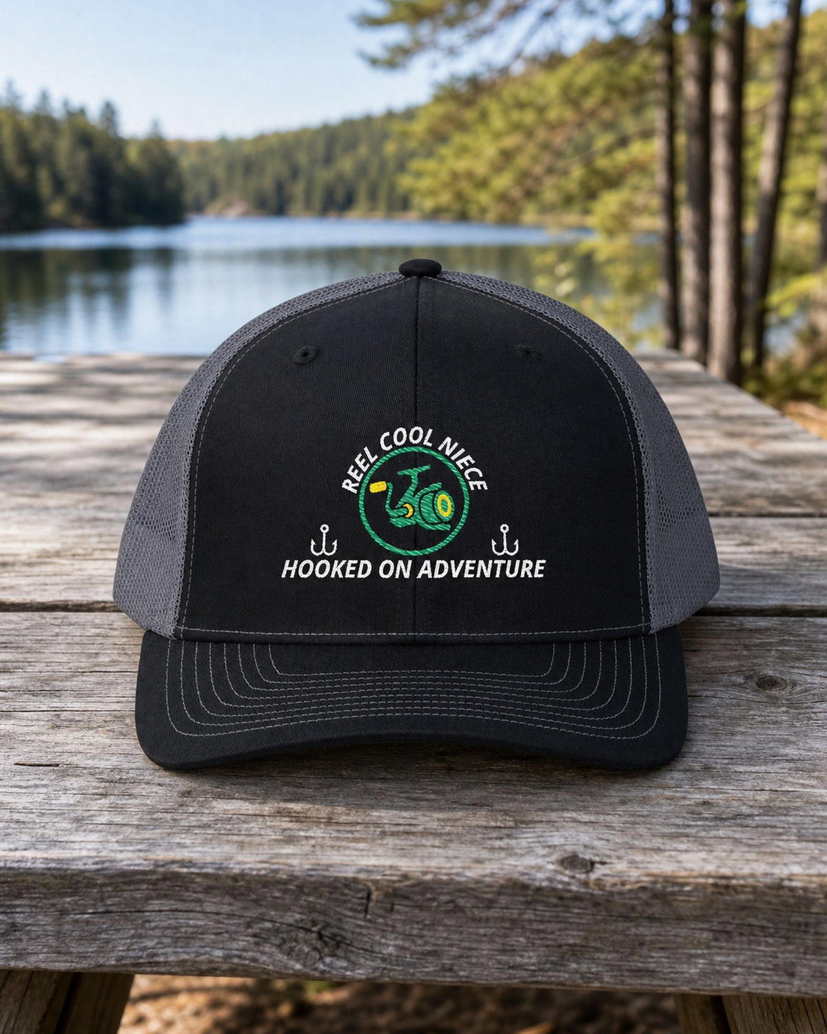 Black and Charcoal REEL COOL NIECE embroidered trucker cap on a full-size weathered lakefront picnic table.