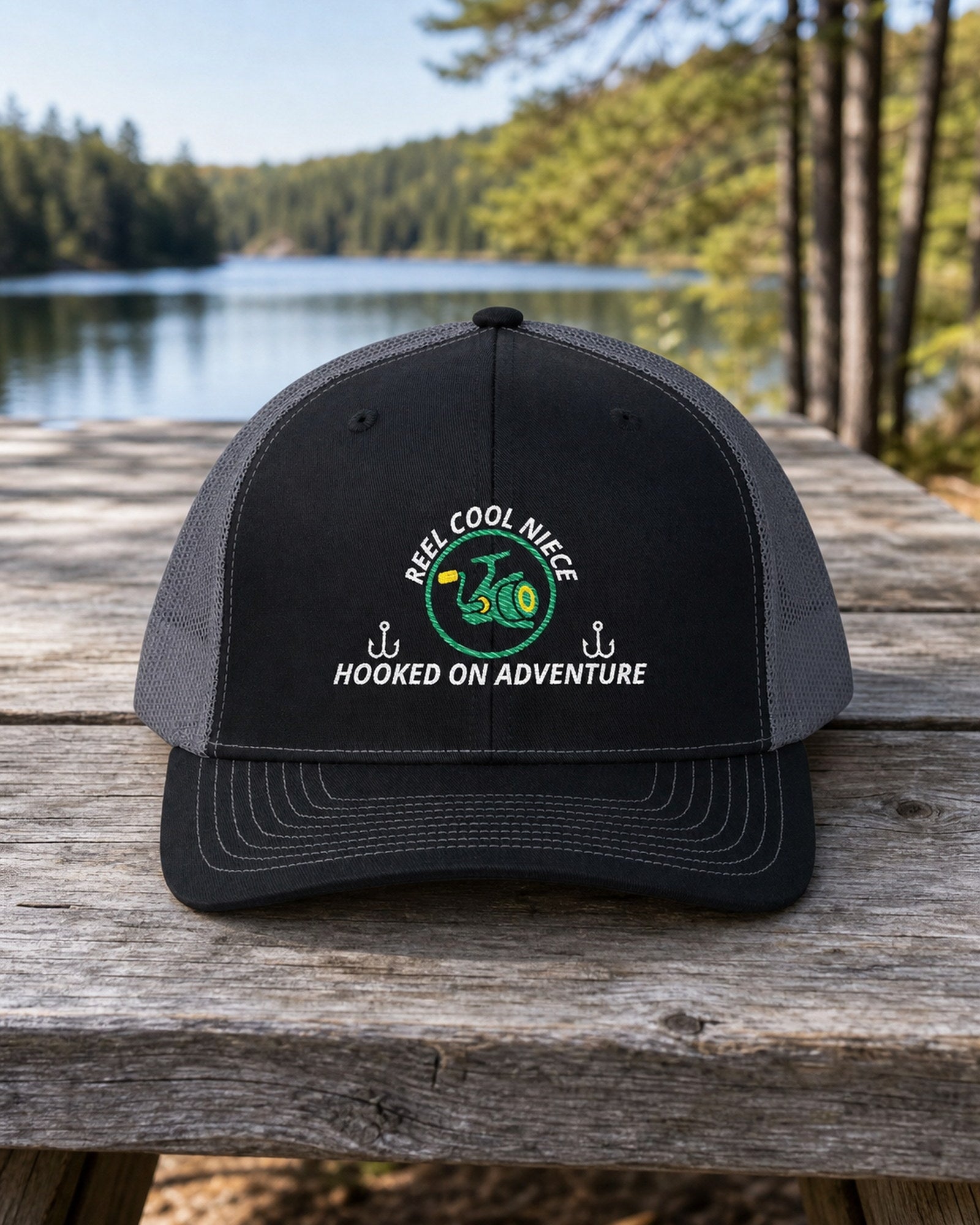 Black and Charcoal REEL COOL NIECE embroidered trucker cap on a full-size weathered lakefront picnic table.