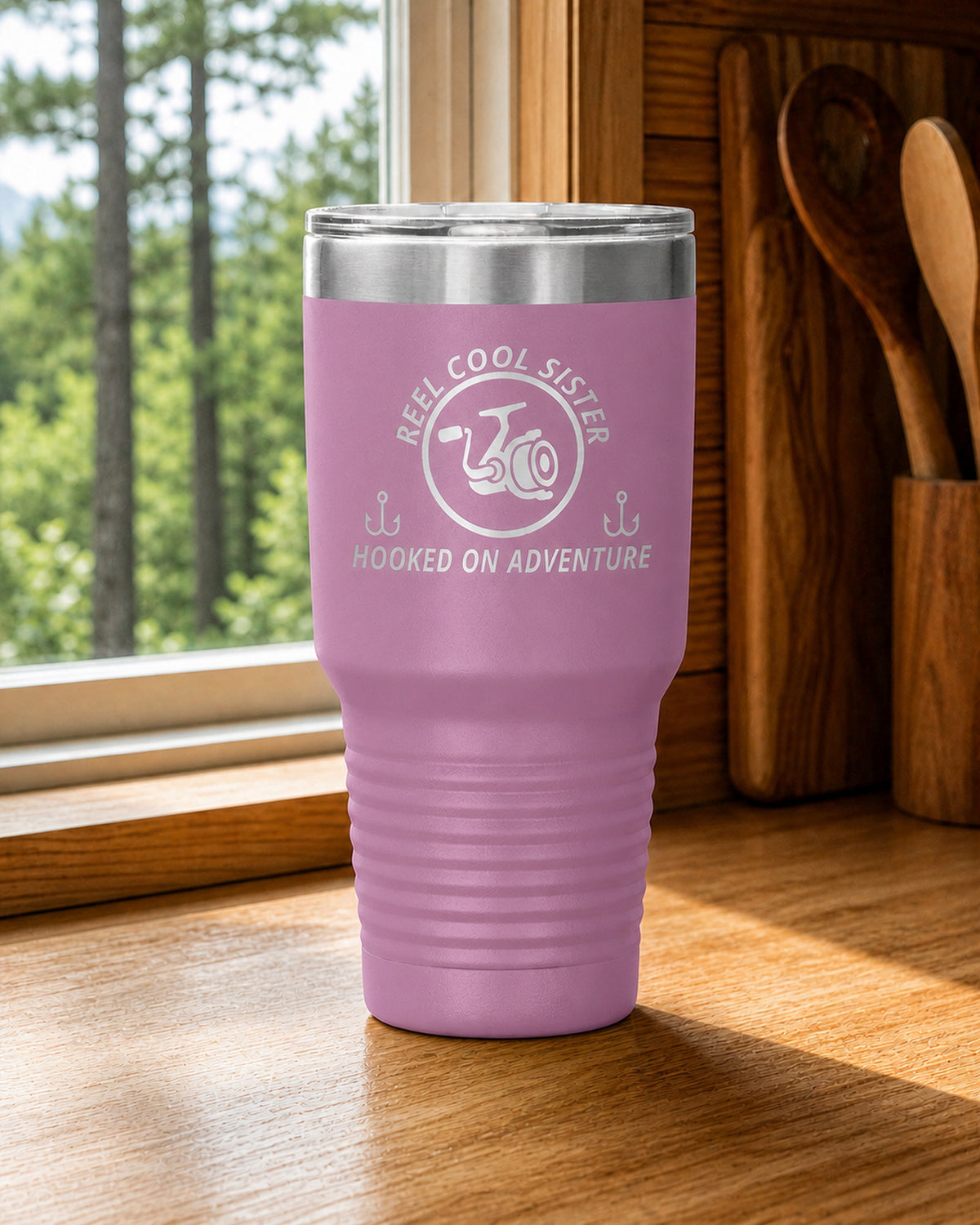 Light Purple 30-ounce REEL COOL SISTER engraved tumbler on a bright wood cabin counter beside a window overlooking green forest.