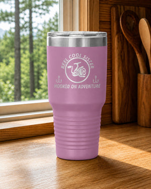 Light Purple 30-ounce REEL COOL SISTER engraved tumbler on a bright wood cabin counter beside a window overlooking green forest.