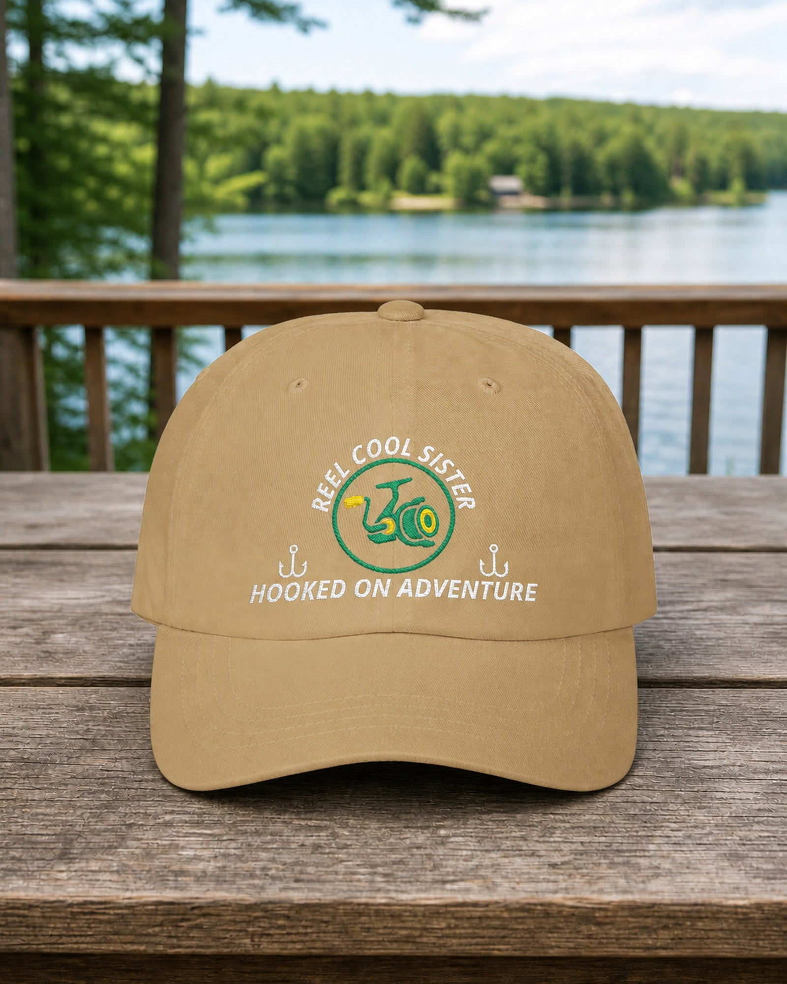 Khaki REEL COOL SISTER embroidered dad cap on a full-size cabin porch table overlooking a pine-lined forest lake
