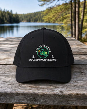Black REEL COOL SISTER embroidered trucker cap on a weathered wooden picnic table beside a pine-lined forest lake.