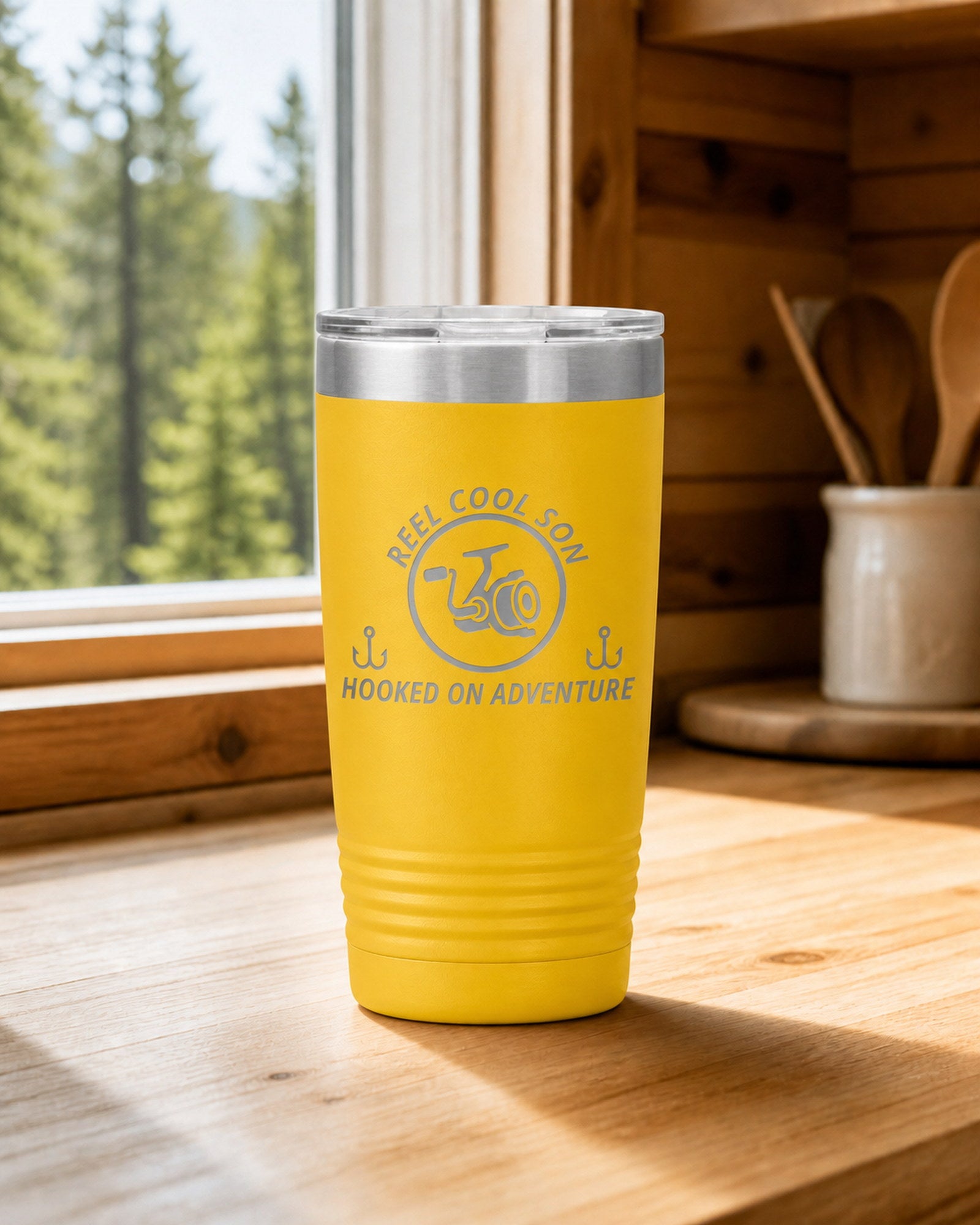 Yellow 20-ounce REEL COOL SON engraved tumbler on a bright wood cabin counter beside a window overlooking green forest.