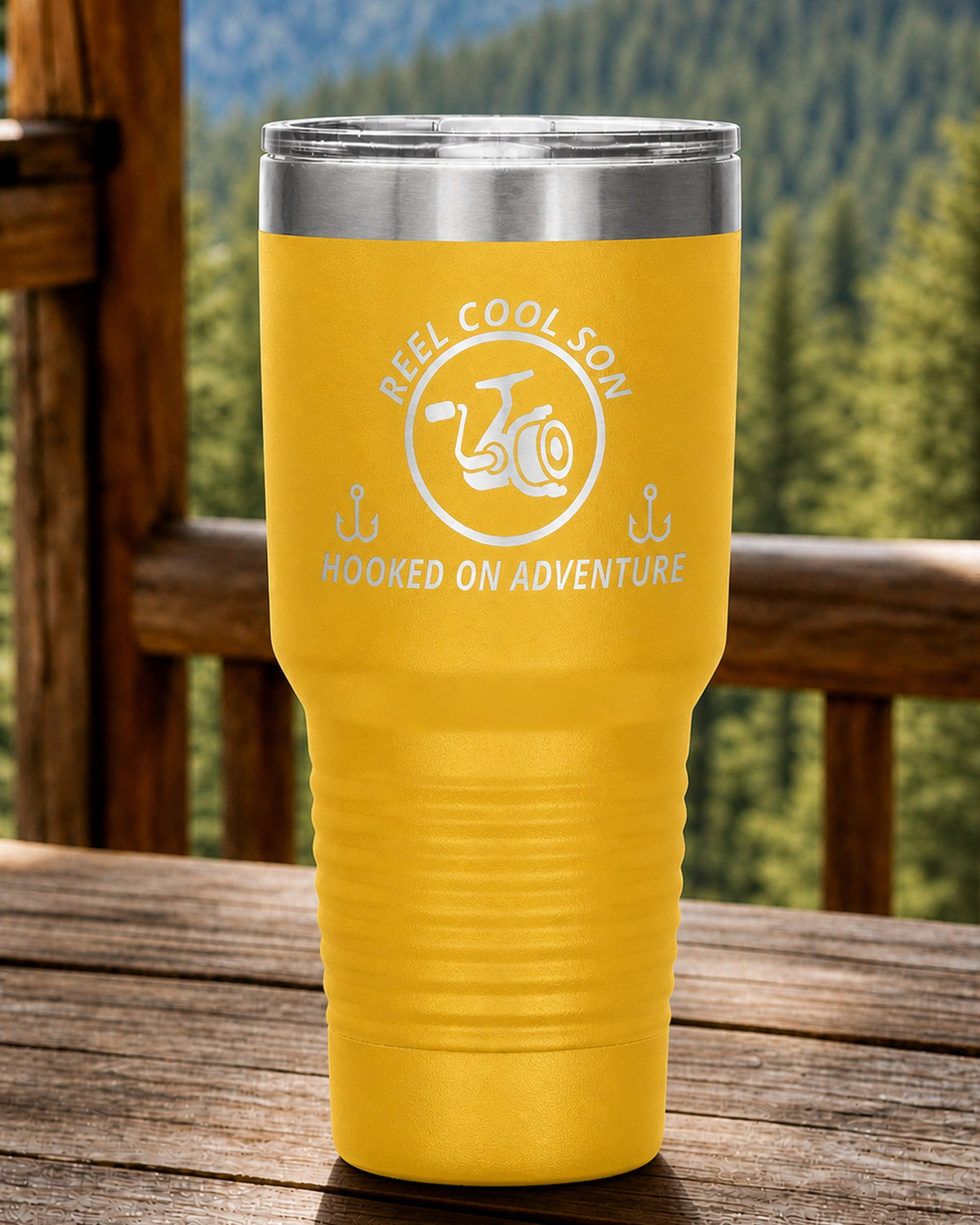 Yellow 30-ounce REEL COOL SON engraved tumbler on a sturdy cabin porch table overlooking forested mountains.