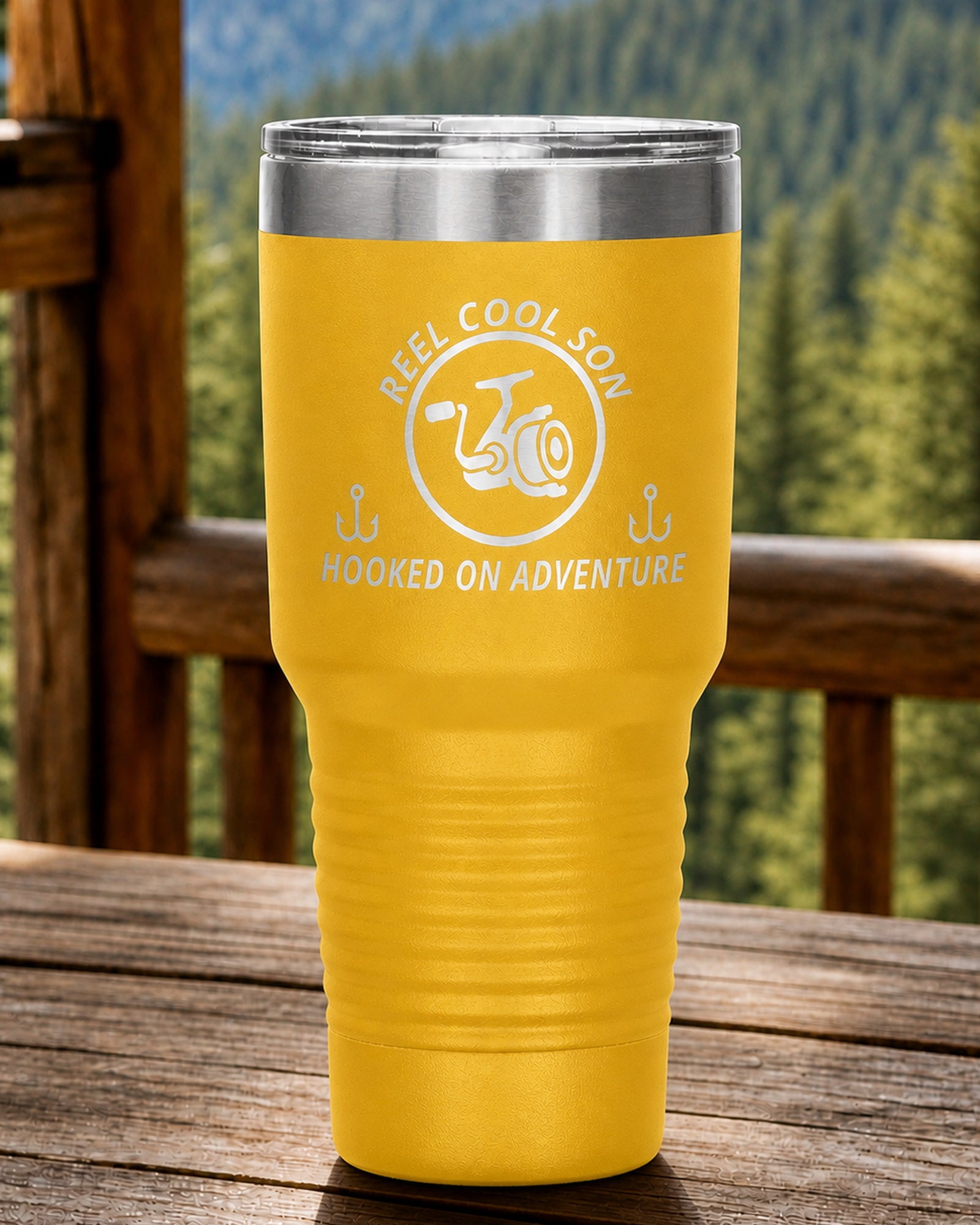 Yellow 30-ounce REEL COOL SON engraved tumbler on a sturdy cabin porch table overlooking forested mountains.