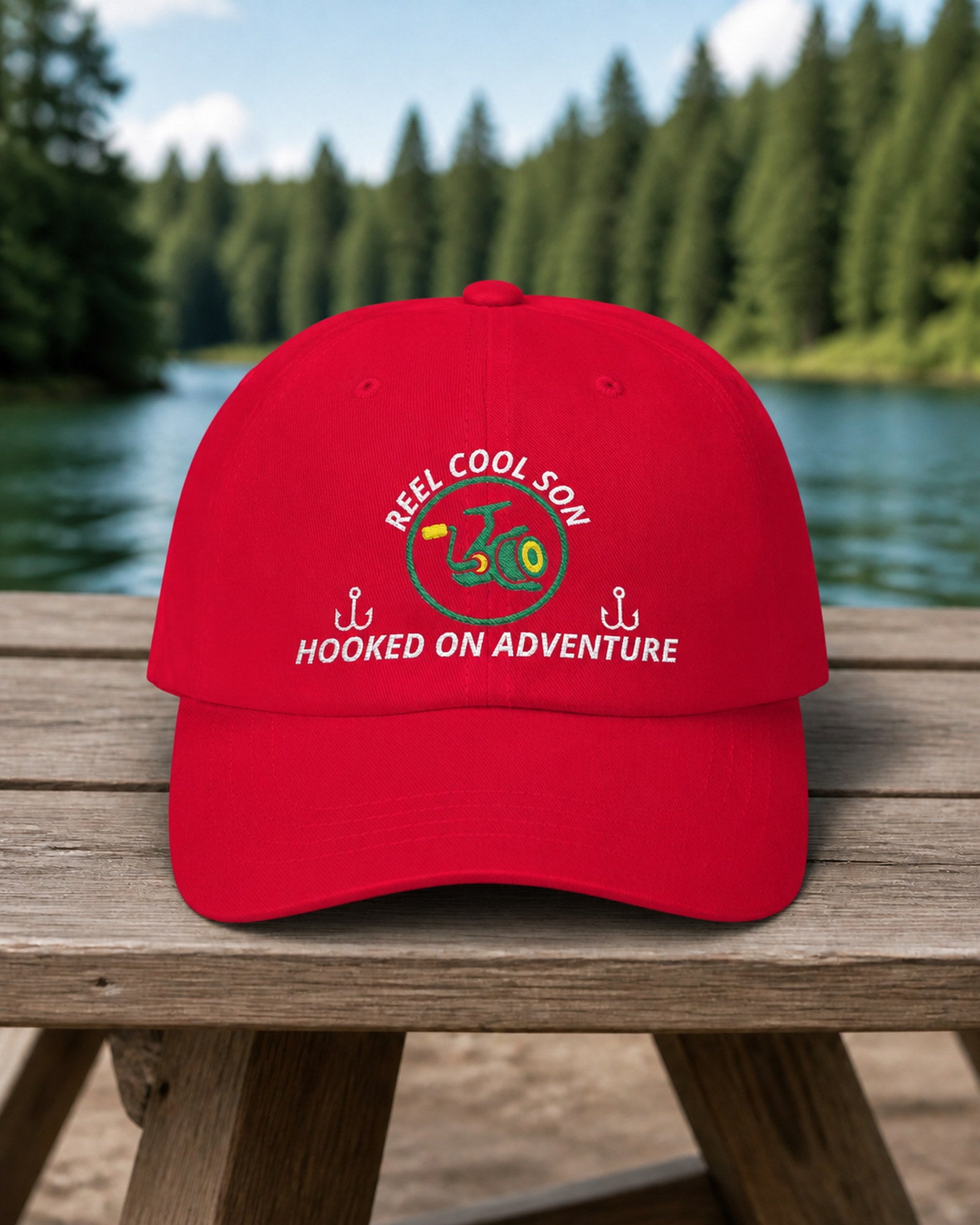 Cranberry REEL COOL SON embroidered dad cap on a weathered wooden picnic table beside a pine-lined forest lake.