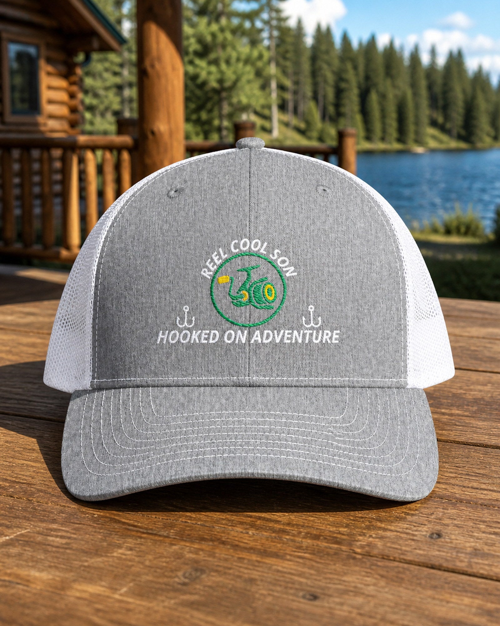 Heather Grey and White REEL COOL SON embroidered trucker cap on a full-size cabin porch table overlooking a pine-lined forest lake.