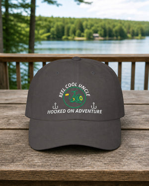 Dark Grey REEL COOL UNCLE embroidered dad cap on a full-size cabin porch table overlooking a pine-lined forest lake.