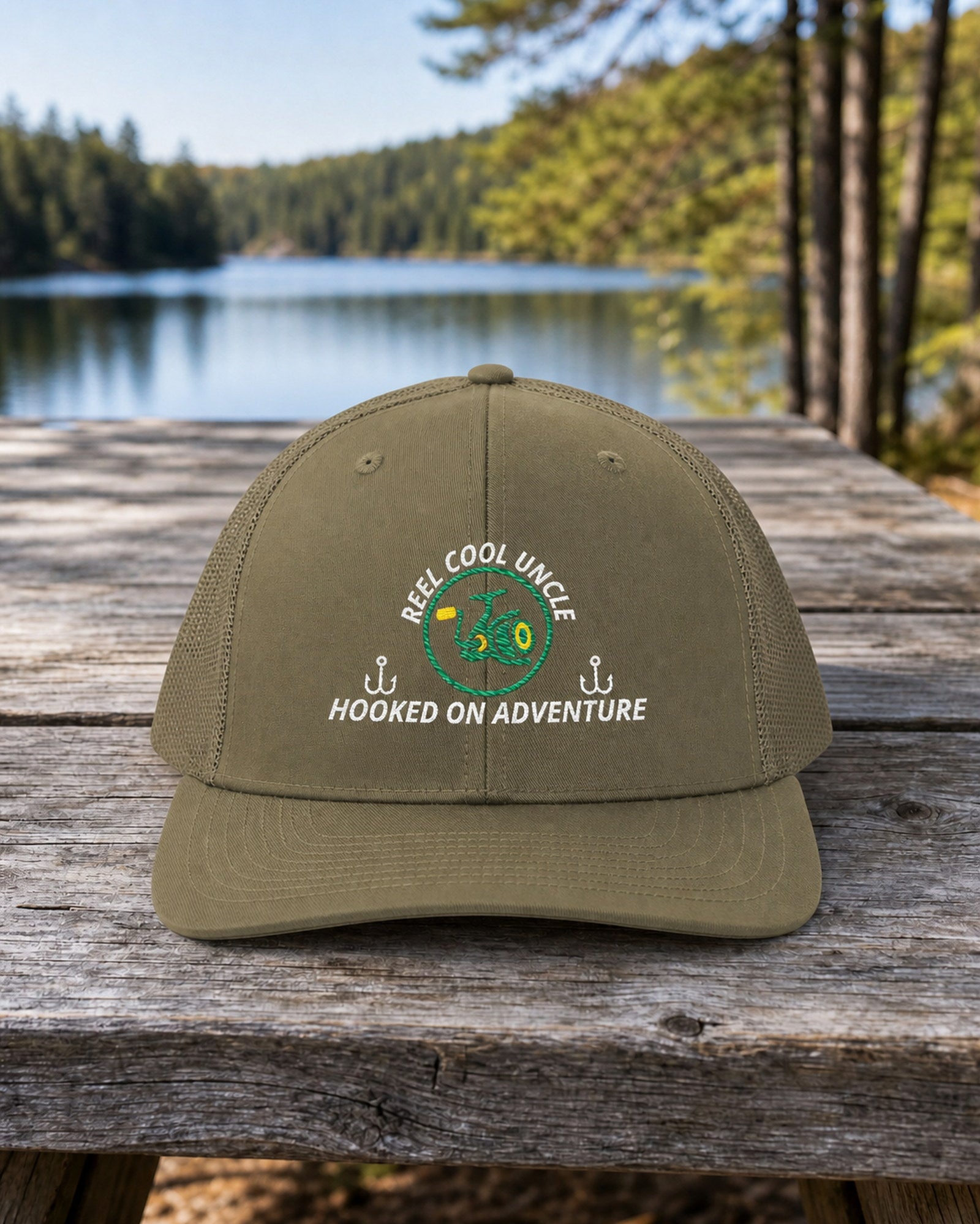 Loden REEL COOL UNCLE embroidered trucker cap on a weathered wooden picnic table beside a pine-lined forest lake.