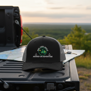 REEL COOL UNCLE trucker cap with black front and charcoal mesh back displayed on a scenic outdoor tailgate.