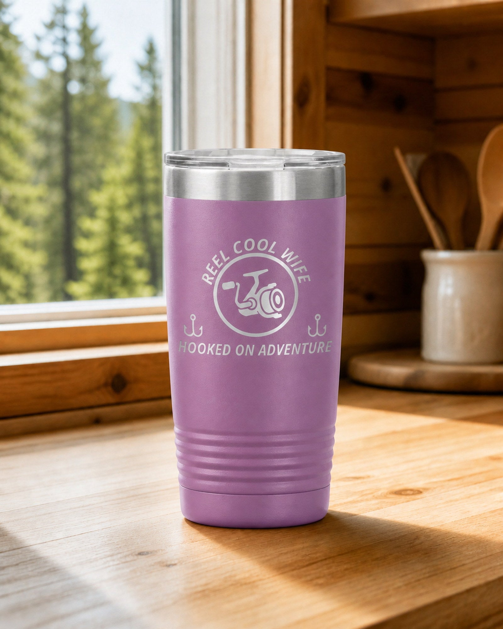 Light Purple 20-ounce REEL COOL WIFE engraved tumbler on a bright wood cabin counter beside a window overlooking green forest.