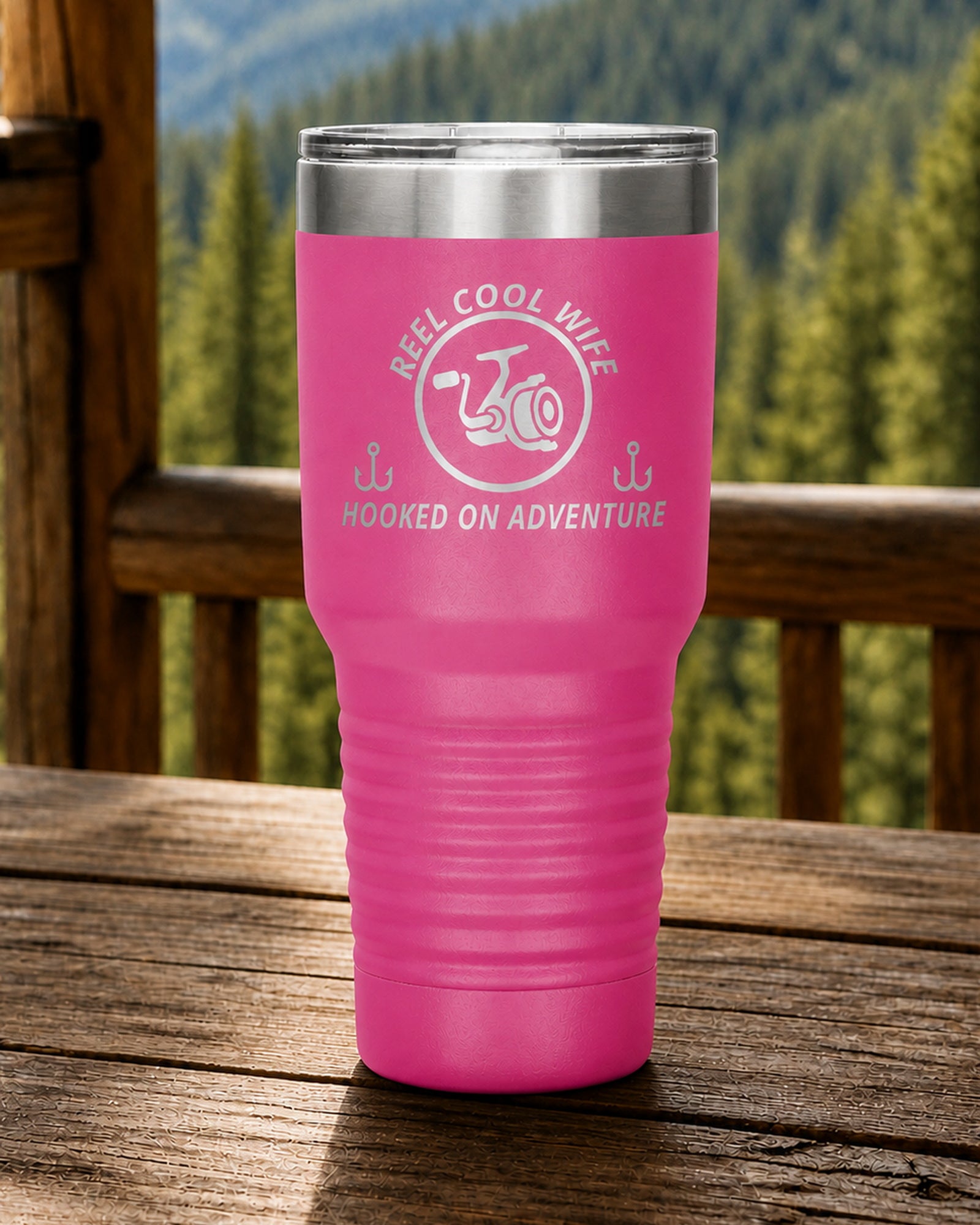 Pink 30-ounce REEL COOL WIFE engraved tumbler on a sturdy cabin porch table overlooking forested mountains.