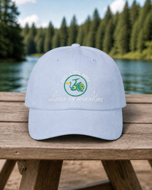Light Blue REEL COOL WIFE embroidered dad cap on a weathered wooden picnic table beside a pine-lined forest lake.