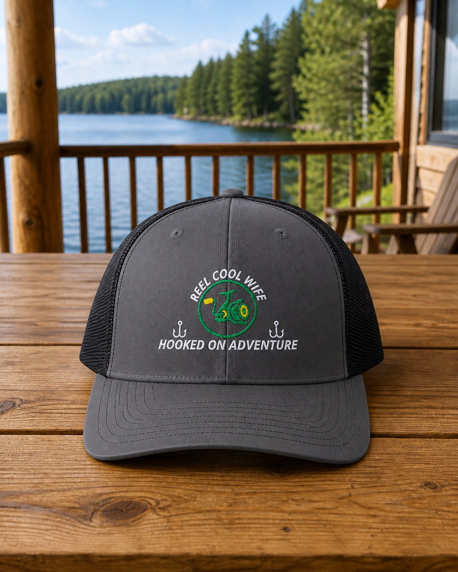 Charcoal and Black REEL COOL WIFE embroidered trucker cap on a full-size cabin porch table overlooking a pine-lined forest lake.